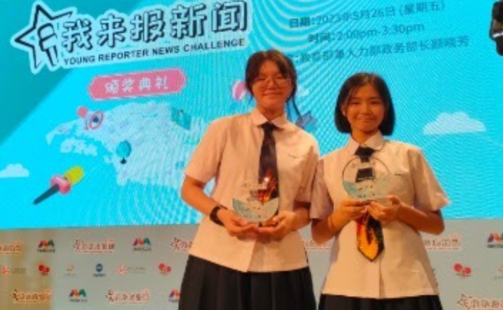 Two young female students in school uniforms hold crystal trophies at a Young Reporter News Challenge award ceremony.